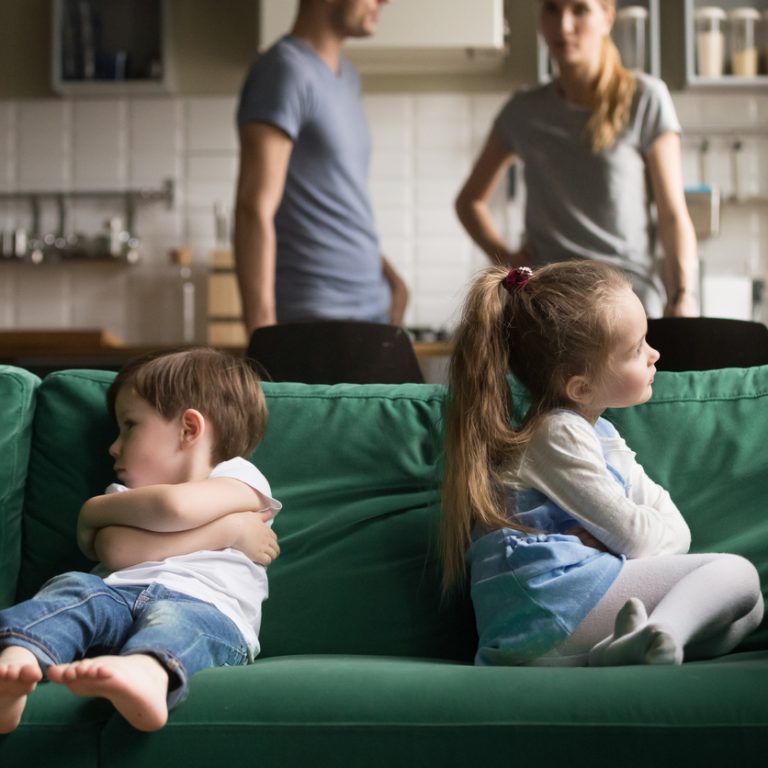 Upset offended toddler brother and sister sitting separately on couch, sofa with arms crossed, little girl and boy ignoring each other, not talking, puzzled parents discuss situation