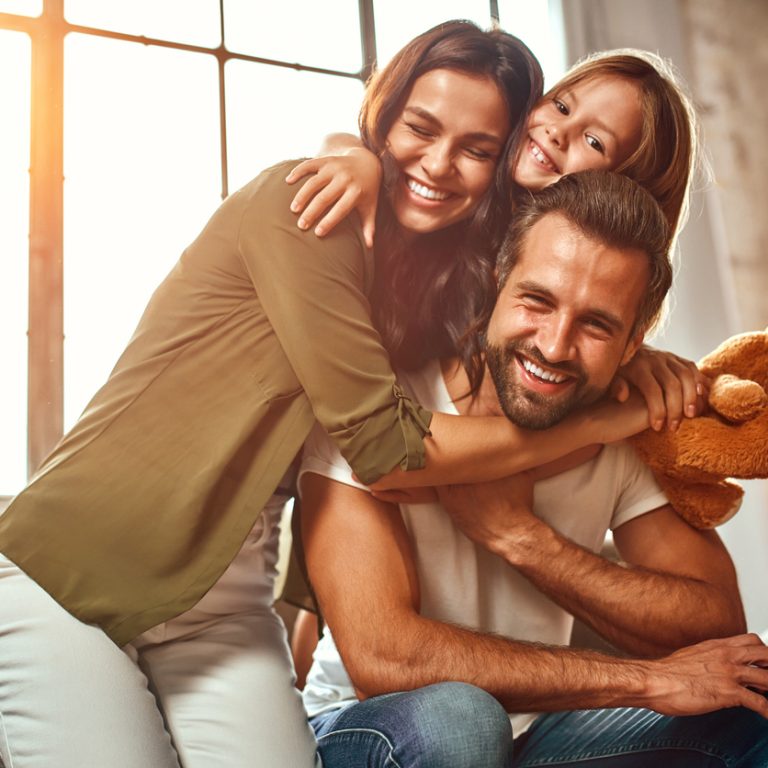 Happy dad and mom with their cute daughter and teddy bear hug and have fun sitting on the sofa in the living room at home.