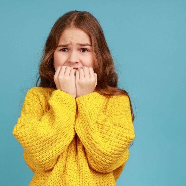 Little girl biting nails with scared big eyes, feeling worried, anxious about childish mistake, wearing yellow casual style sweater. Indoor studio shot isolated on blue background.