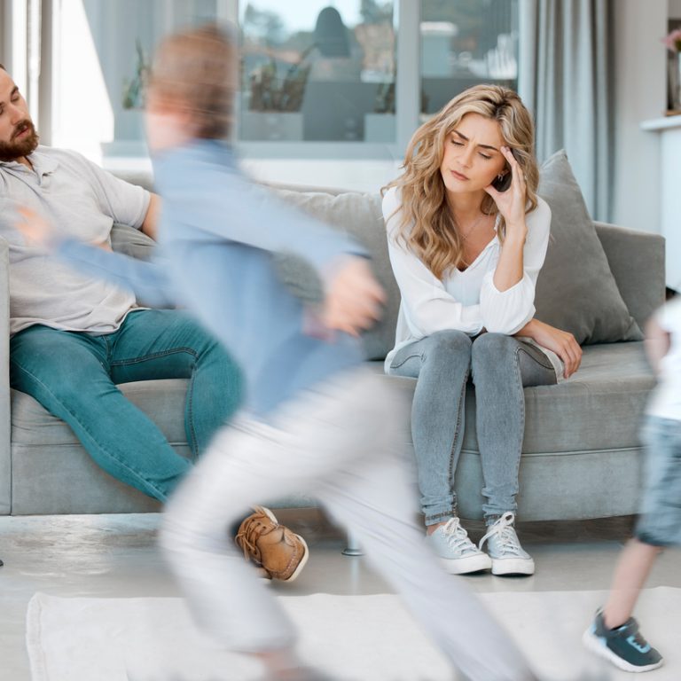 Unhappy young caucasian couple sitting on the sofa looking stressed and depressed while their hyper naughty little children play around them in the living room at home in a bright room